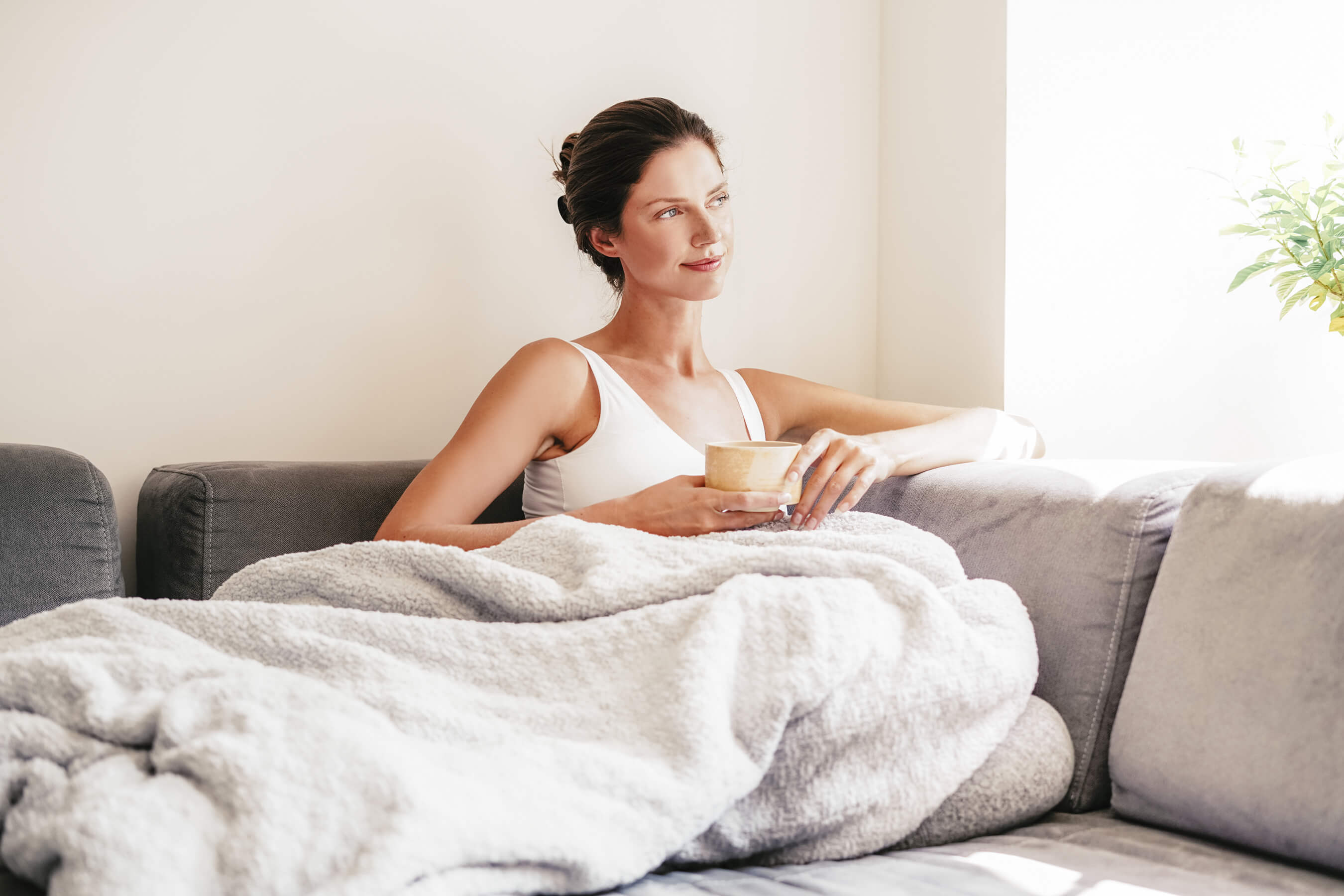 Woman sitting on the couch with a weighted blanket. Sunday Citizen Snug Crystal Weighted Blanket. Woman relaxing with weighted blanket.