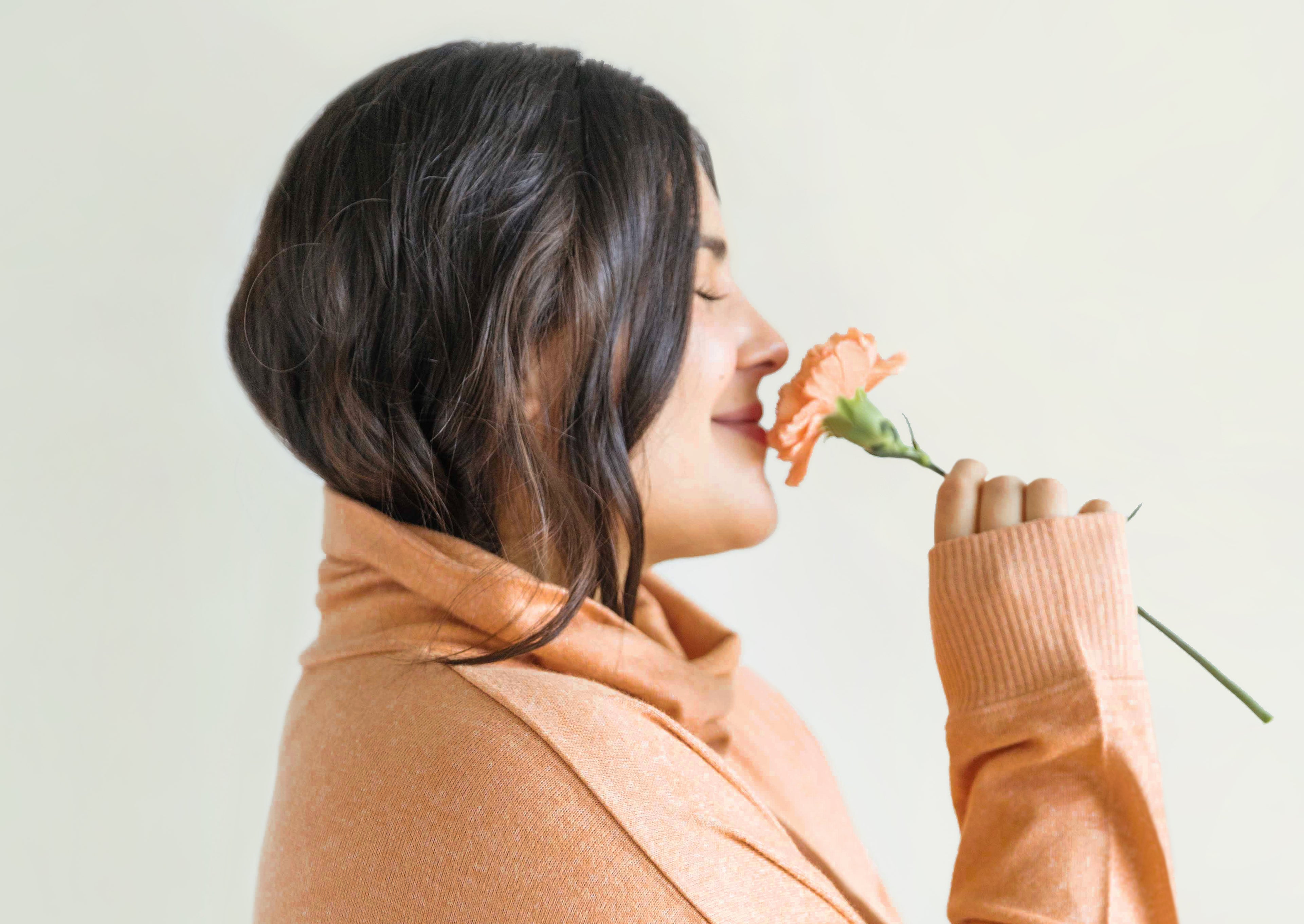 Woman sniffing flower. Woman wearing orange turtleneck and sniffing orange flower. Woman wearing Sunday Citizen Cosset Relaxed Turtleneck in Camel.