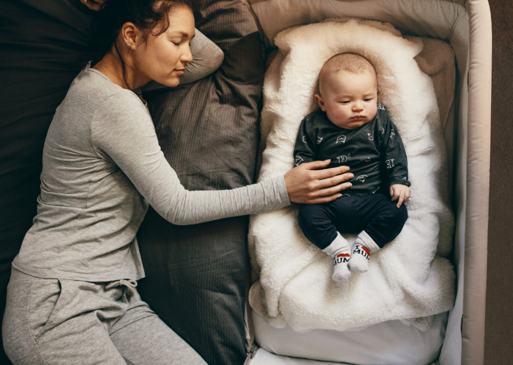 A mother sleeping next to a baby in a bassinet.