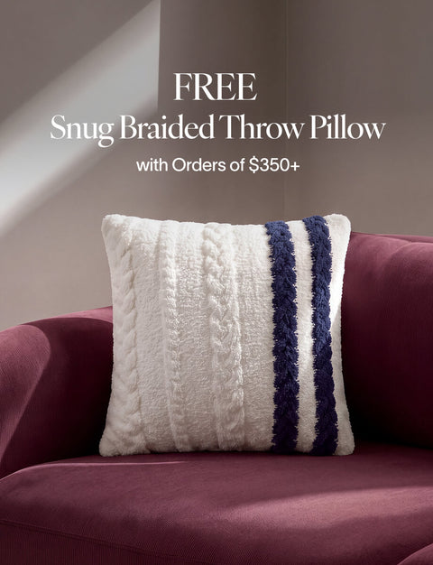 A cream cable-knit throw pillow with two vertical navy blue stripes sits on a burgundy velvet sofa. Text at the top reads 'FREE Snug Braided Throw Pillow with Orders of $350+'.