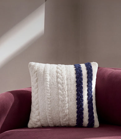 A cream cable-knit throw pillow with navy blue stripes, resting on a burgundy velvet sofa against a neutral beige wall.