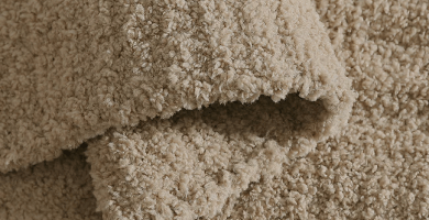 Close-up of a beige shaggy or looped carpet texture with a folded corner showing the pile direction.