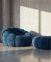Two modern, cloud-like bouclé poufs in a deep teal-blue color, styled in a minimalist concrete room with dramatic sunlight and shadows.