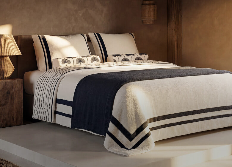 A styled bedroom scene showing a bed dressed in white bedding with dark navy stripes and a navy throw blanket, set in a warm-toned interior with bedside tables, lamps, and a window.