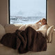 Sunday Citizen Snug Knit Chunky Throw in Chocolate Martini draped over a person on a sofa with a snowy mountain view